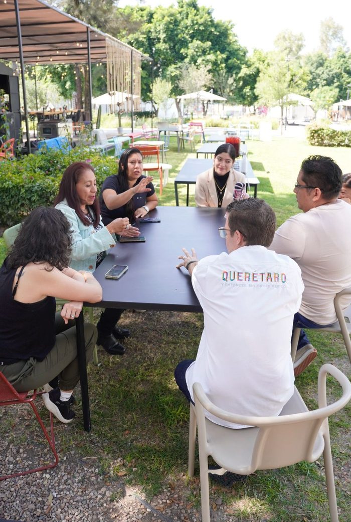 Grupo de personas conversando alrededor de una mesa al aire libre, una de ellas con una playera de Querétaro